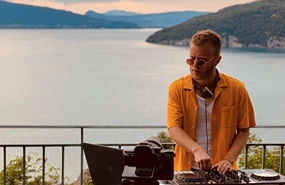 Un DJ qui mix sur des platines lors d'une soirée sunset couché de soleil vue sur la mer. Il a un casque de musique et une sonnorisation de DJ